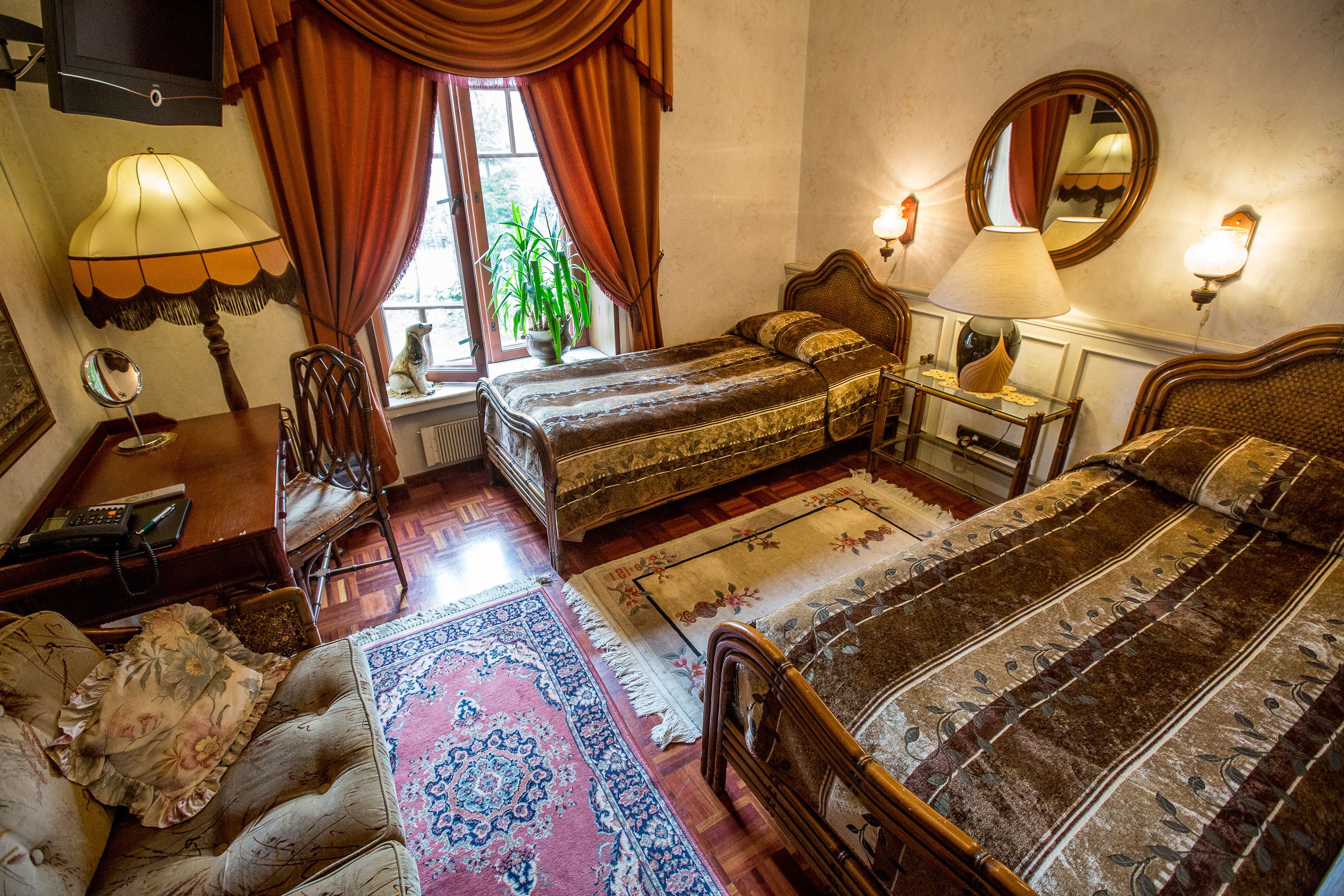 Un dormitorio tradicional con dos camas, una zona de estar con sofá, suelo de madera y un gran ventanal con cortinas.