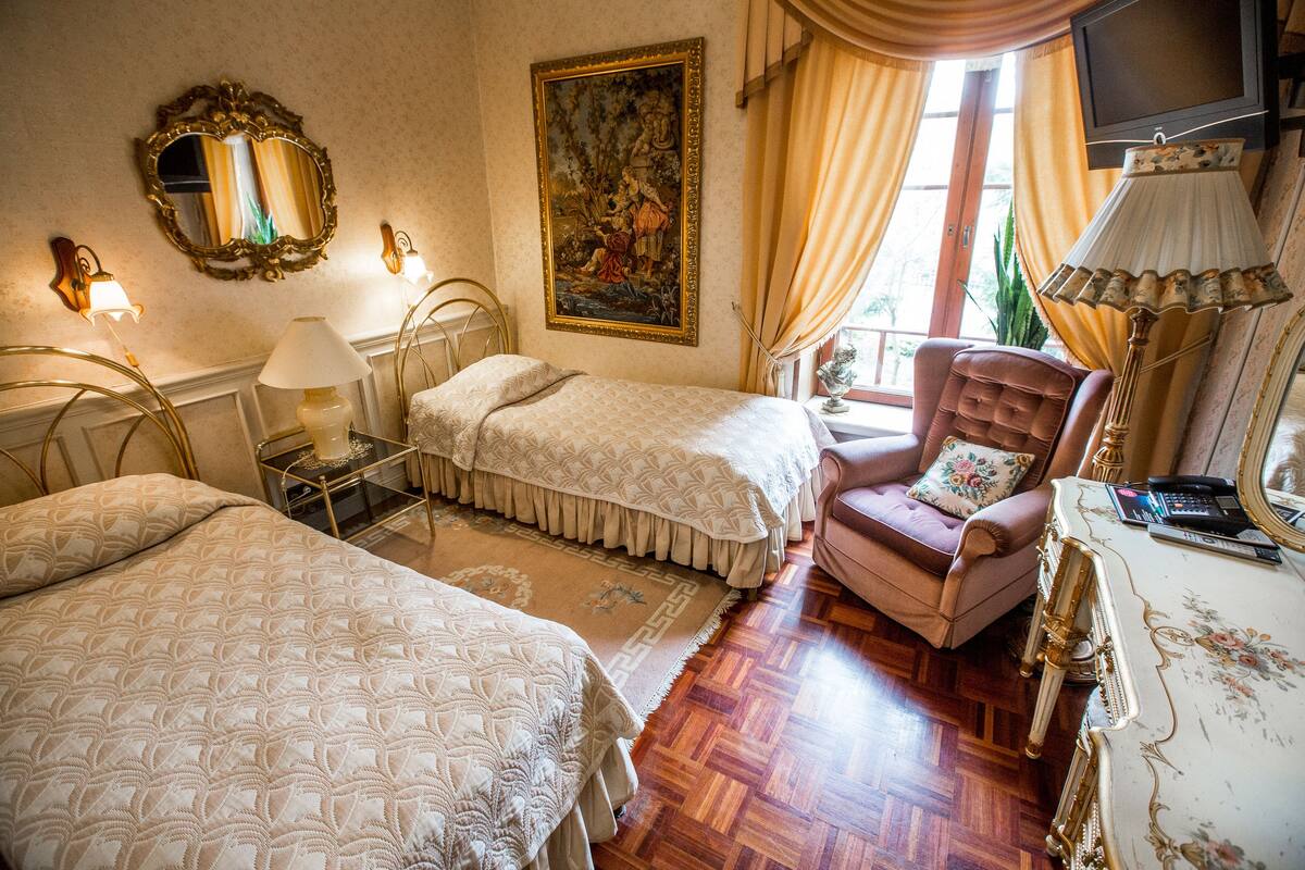 A hotel room with two beds, a sitting area with a chair, a table with a lamp, a painting on the wall, and a television.