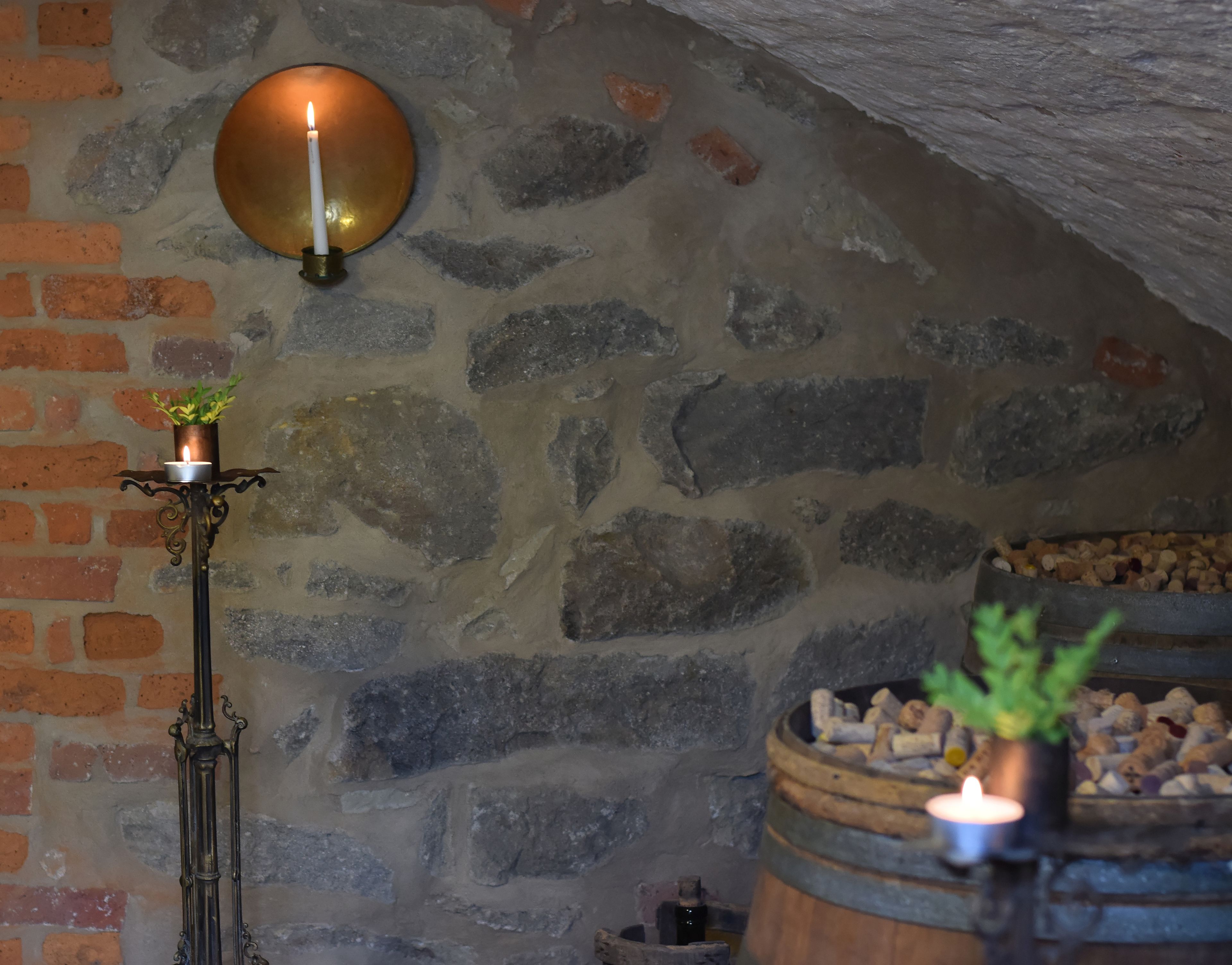 Un muro de piedra con una sección de ladrillo, un candelabro de metal con una vela encendida, y un barril lleno de leña.