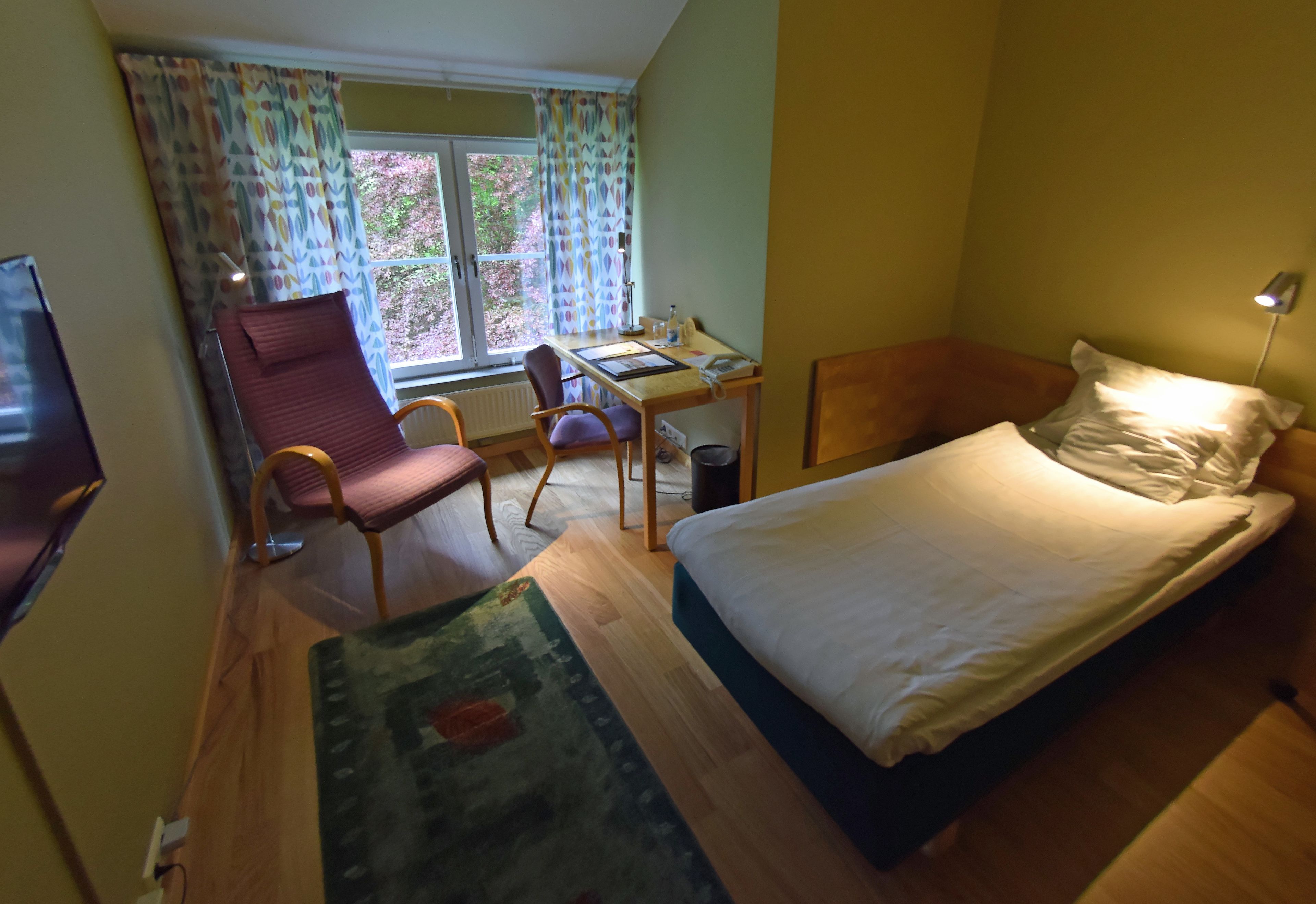 A compact hotel room with a single bed, a desk with a chair, a television, and a window with curtains.