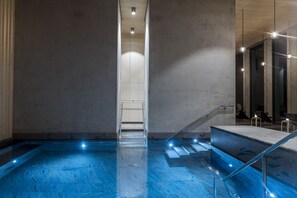 Un espace piscine intérieur moderne, doté de garde-corps en verre transparent, d’un escalier central et d’un éclairage d’ambiance.