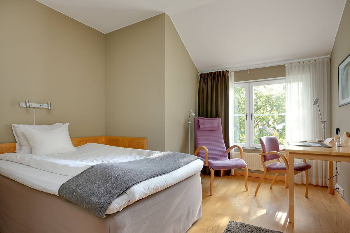 Une chambre d’hôtel avec un lit, un bureau, deux chaises, une fenêtre avec des rideaux et un radiateur.