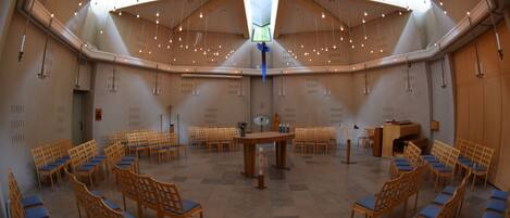 A spacious hall with rows of wooden chairs and tables, a central altar, and a high ceiling with skylights.