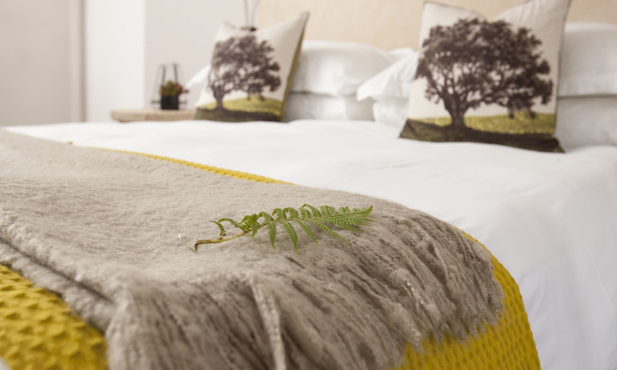 Un lit bien fait, recouvert d’une couverture grise et jaune, avec deux oreillers ornés d’un motif d’arbre et une petite plante posée sur le lit.