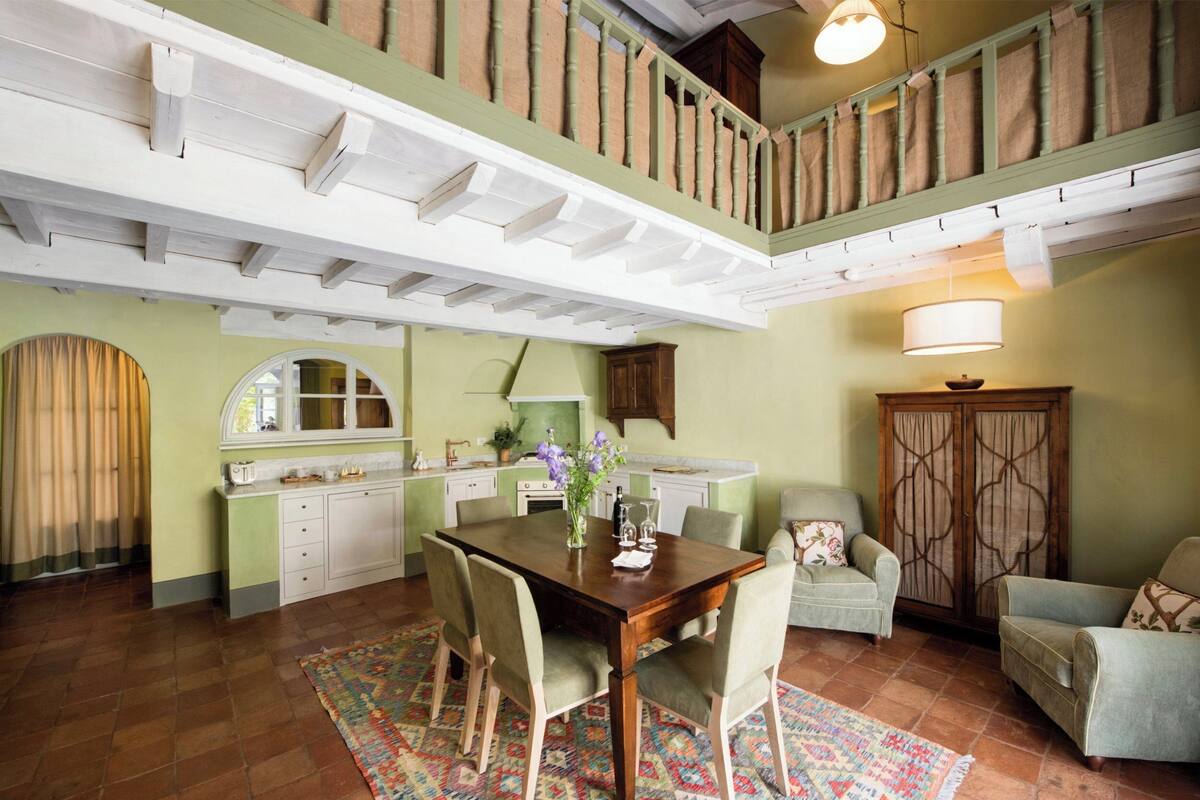 Un comedor con mesa y sillas de madera, una cocina al fondo y una escalera que sube a un nivel superior.