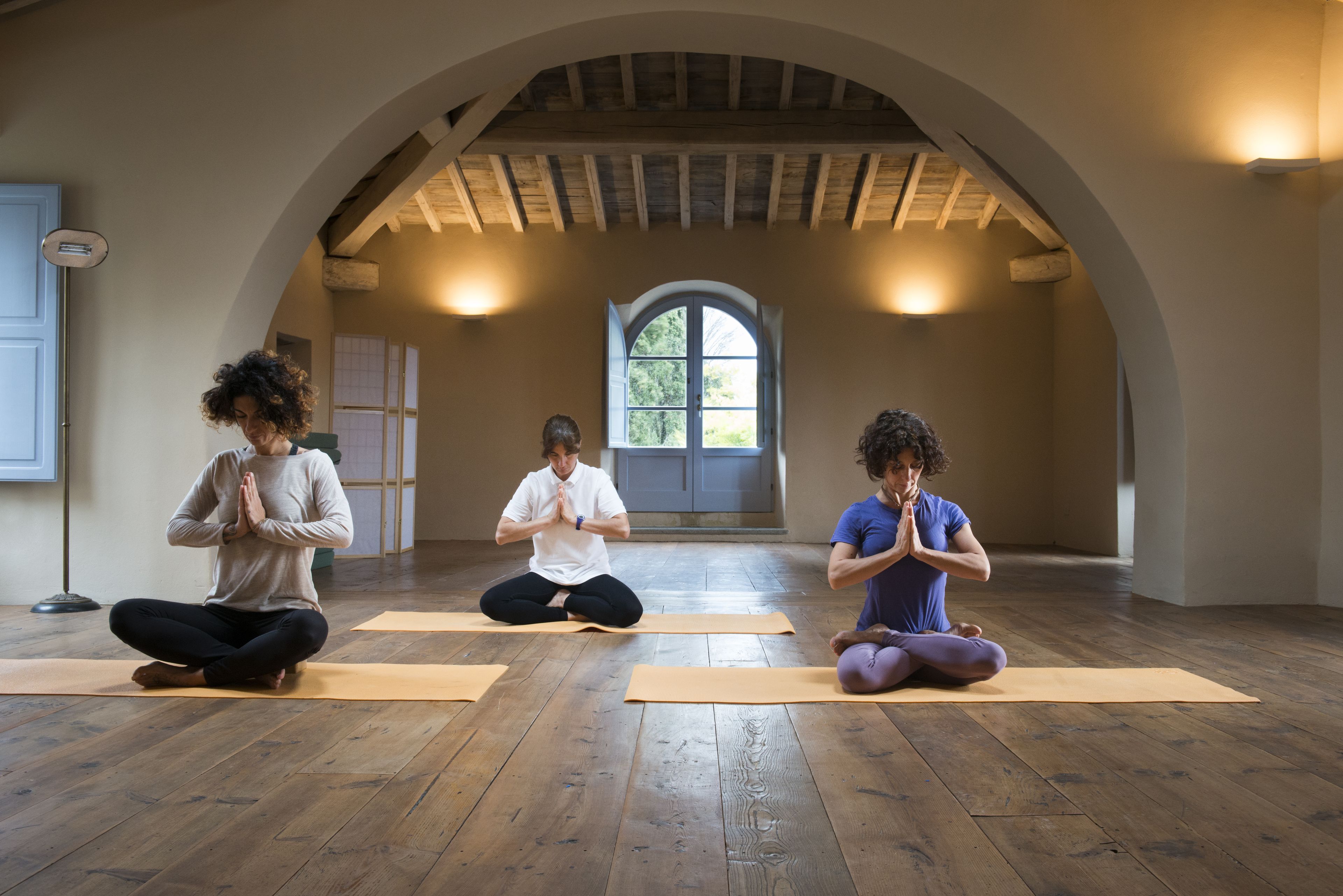 Three people meditating on yoga mats in a room with wooden floors and an arched ceiling.