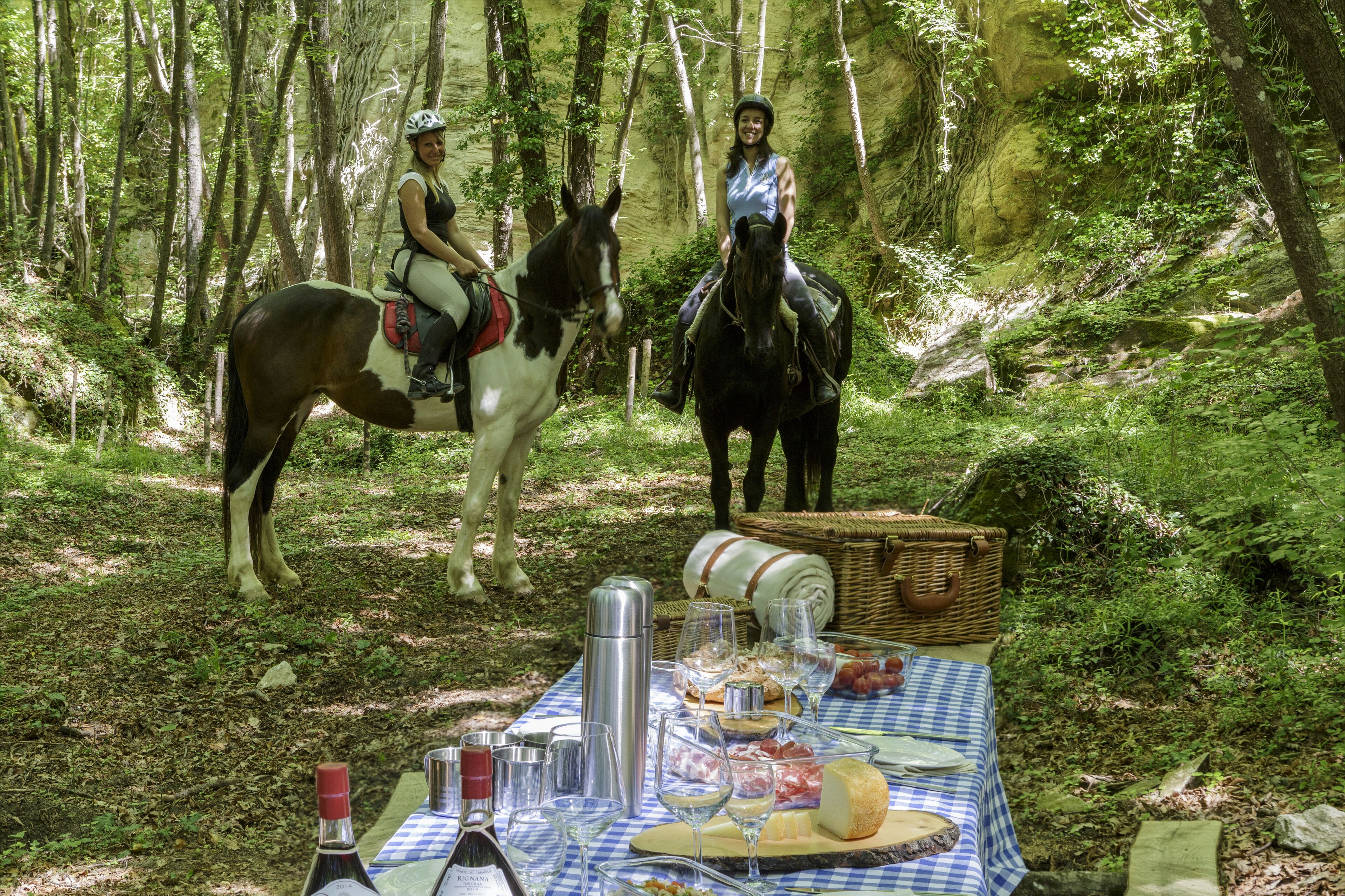 Two people on horseback in a forest with a picnic table set up.