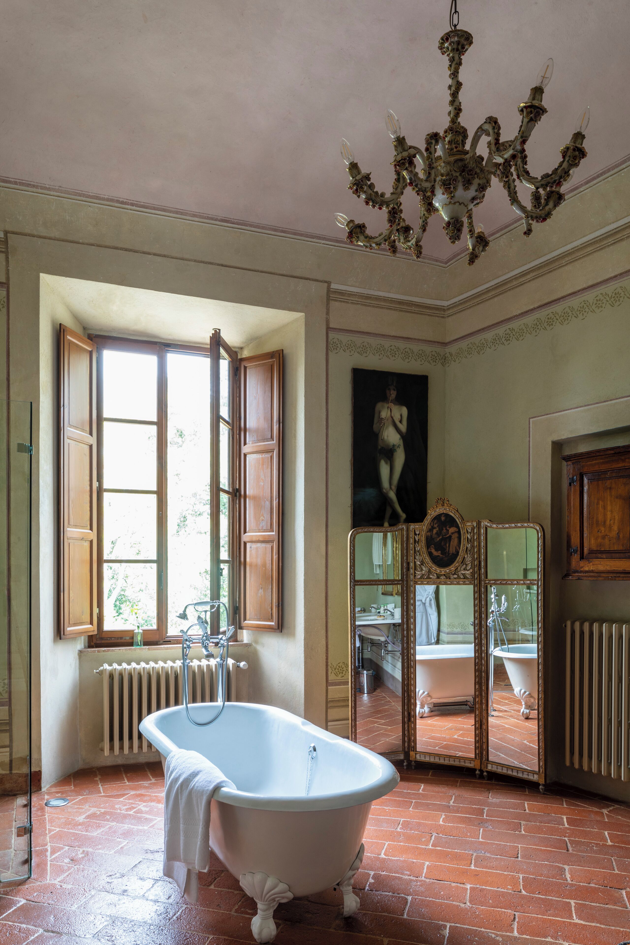Ein Badezimmer mit freistehender Badewanne, einem großen Fenster mit Holzläden, einem Heizkörper und einem Spiegel mit dekorativem Rahmen.