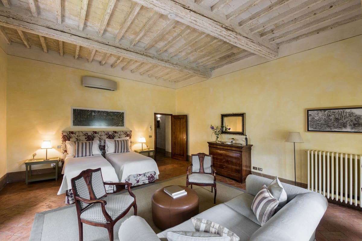 Una habitación de hotel con una cama, dos sillas, un sofá, un aparador de madera, un radiador y un cuadro en la pared.