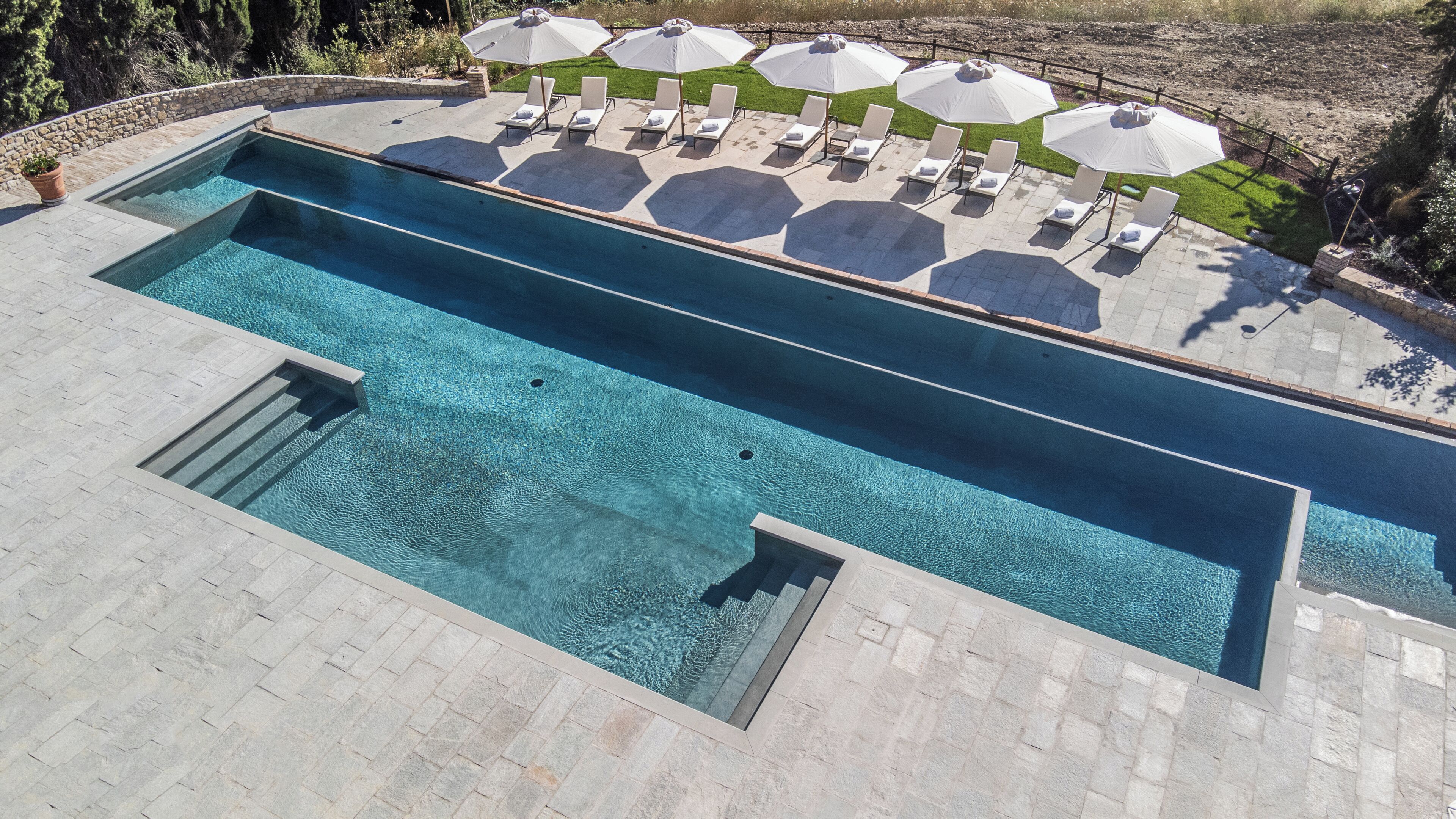 Ein rechteckiger Pool mit einer Steindeck, umgeben von Liegestühlen und Sonnenschirmen.