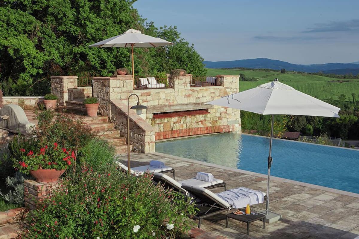 Zona de piscina con sillas de descanso, sombrillas y una pared de piedra.