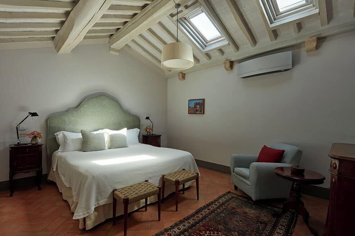 Un dormitorio con una cama grande, una silla, una mesita, una mesilla de noche, una lámpara, un cuadro y un tragaluz.