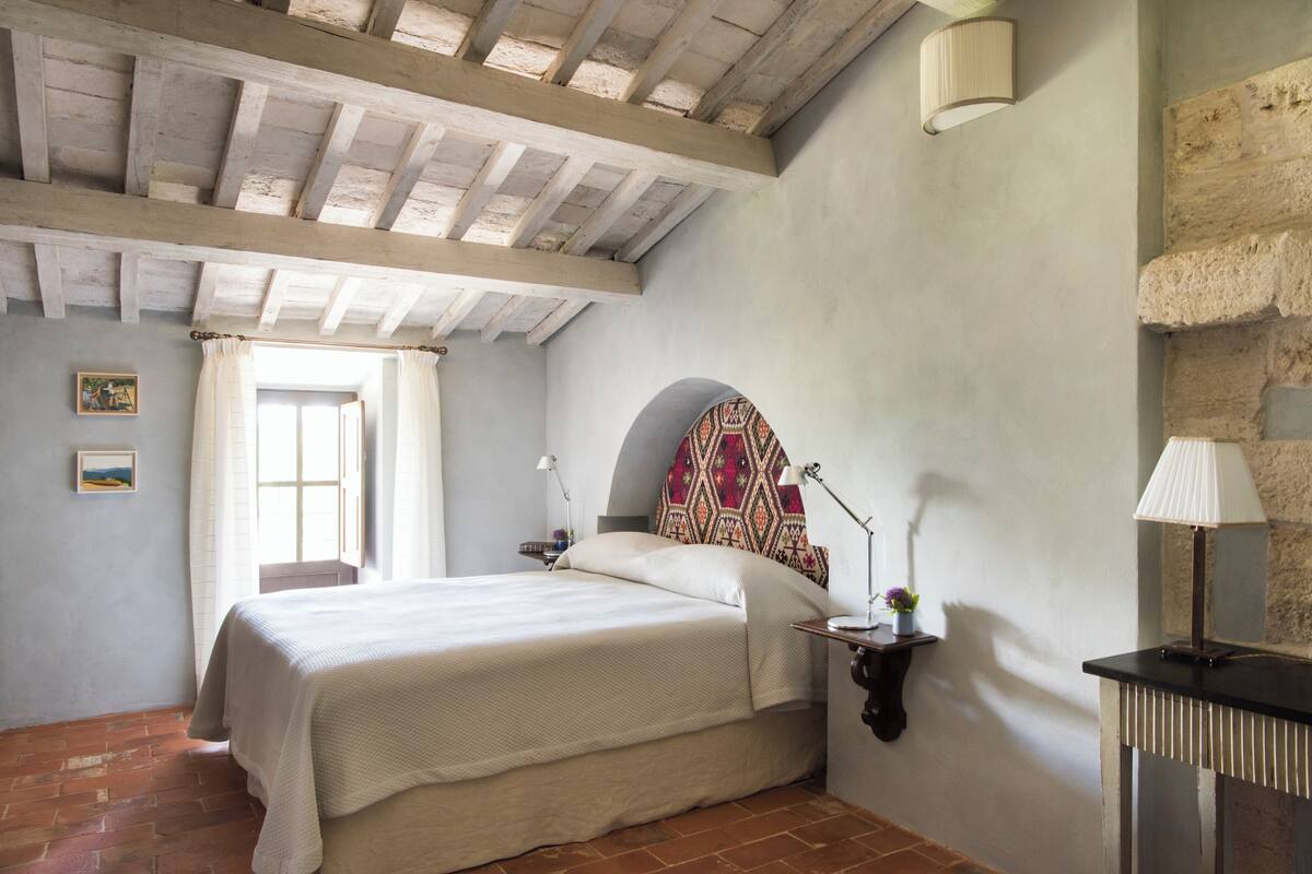 Un dormitorio con cama, mesita de noche, lámpara, ventana con cortinas, lámpara de pared y pared de piedra.