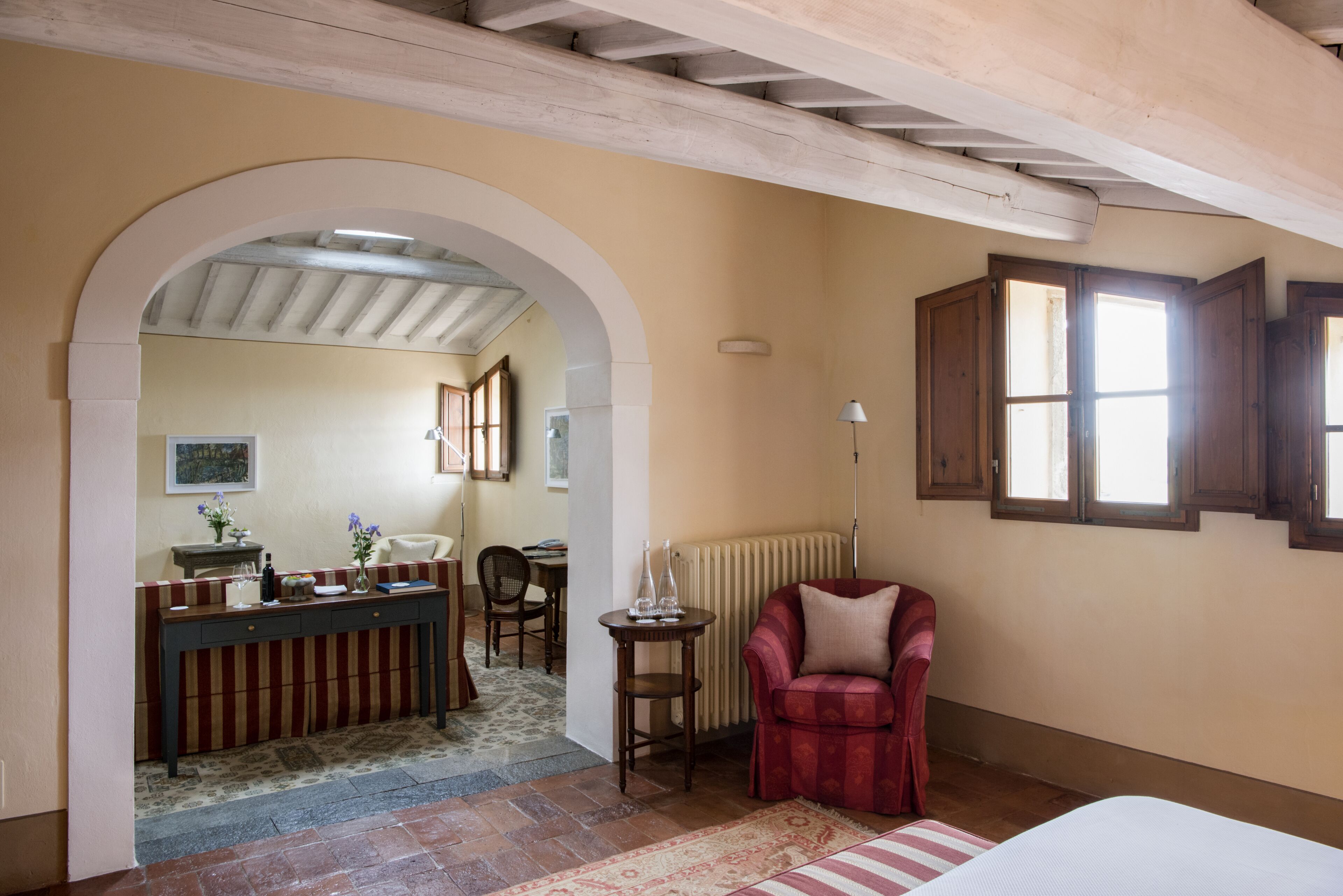 A room with a bed, a red armchair, a wooden desk with a chair, and a window with wooden shutters.