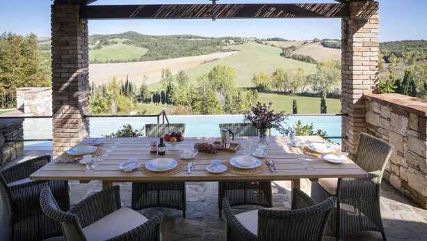 Een overdekt terras met een tafel voor vier personen, met uitzicht op een schilderachtig landschap met heuvels en een waterpartij.