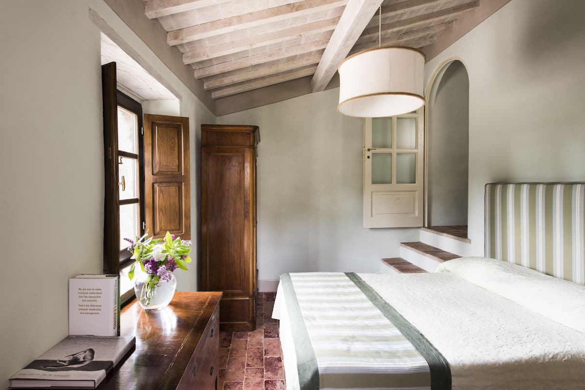 Un dormitorio con un armario de madera, una mesita de noche con un libro y un florero, una cama con cabecera a rayas y una ventana con vistas al exterior.