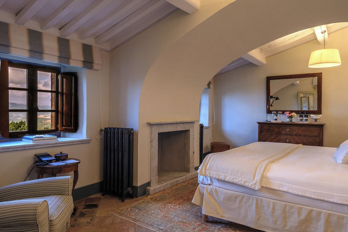 Un dormitorio con cama, chimenea, ventana con vistas, escritorio con laptop y espejo.