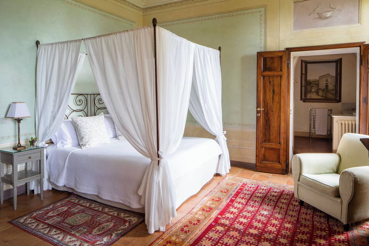 Una cama con dosel y cuatro columnas con ropa blanca, una mesita de noche con lámpara y una alfombra roja estampada.