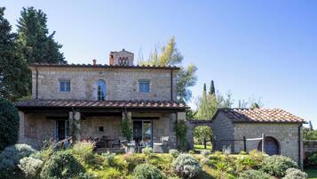 Une maison en pierre avec un toit en tuiles, une terrasse couverte avec des chaises et un jardin où poussent différentes espèces de plantes.