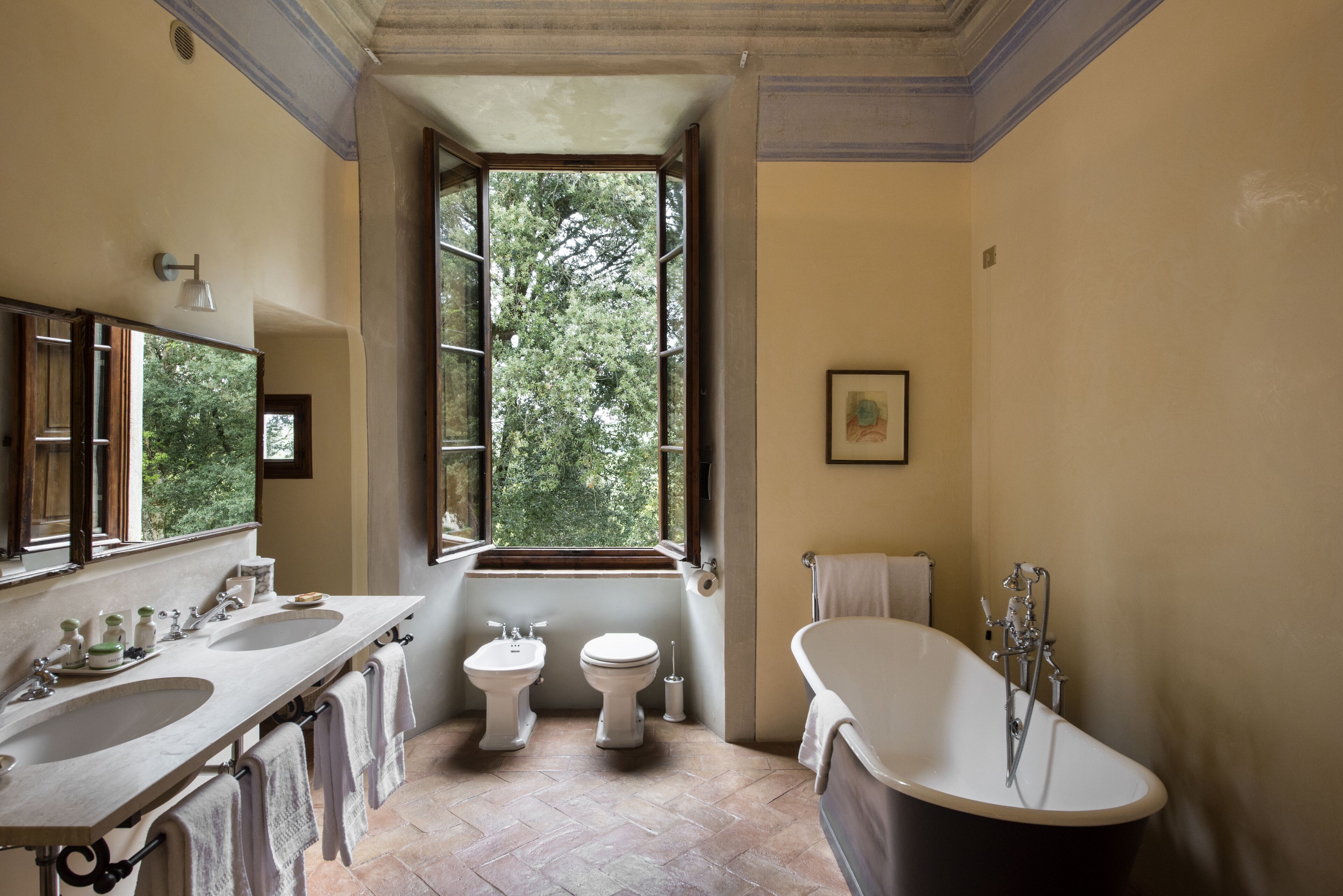 A bathroom with two sinks, a toilet, and a bathtub. There is a window providing natural light and a view of trees.
