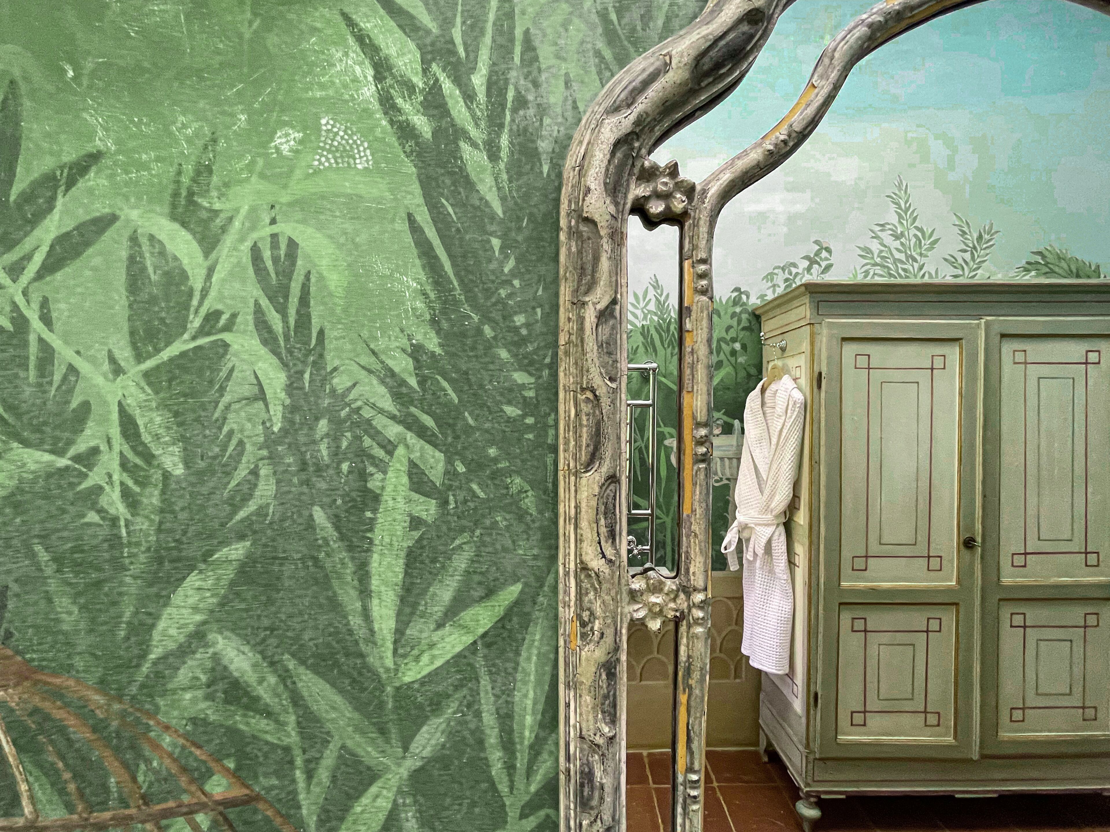 Une salle de bain ornée d’une fresque murale représentant des feuilles vertes, d’un miroir encadré en bois et d’un meuble sur lequel est accrochée une serviette.
