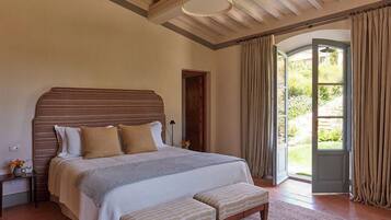 A bedroom with a large bed, a bench, a nightstand, a lamp, and a view of the outdoors through a door.
