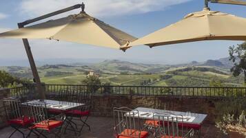 A terrace with tables and chairs, a view of rolling hills, and a stone wall.