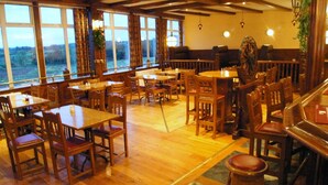 A restaurant with wooden tables and chairs, large windows with curtains, and a view of the outdoors. - The Hedges Hotel (Ballymoney)