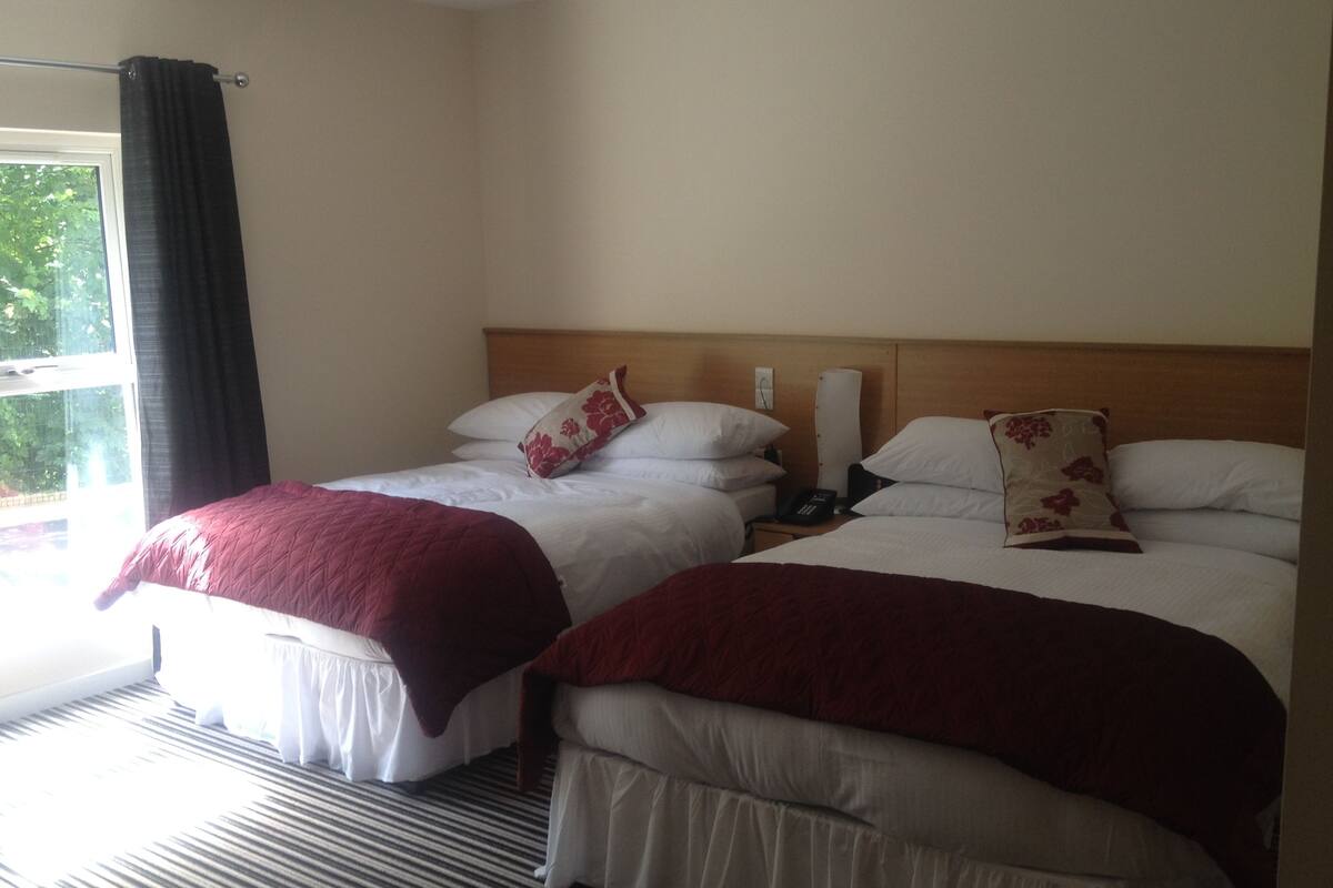 A hotel room with two beds, each with white bedding and red throw pillows, a wooden headboard, a bedside table with a lamp, and a window with curtains.
