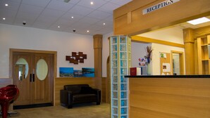 A reception area with a wooden reception desk, a black leather couch, and a red chair. - The Hedges Hotel (Ballymoney)
