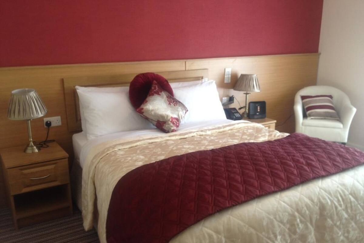 A hotel room with a bed, two bedside tables, a lamp, a chair, and a red wall.