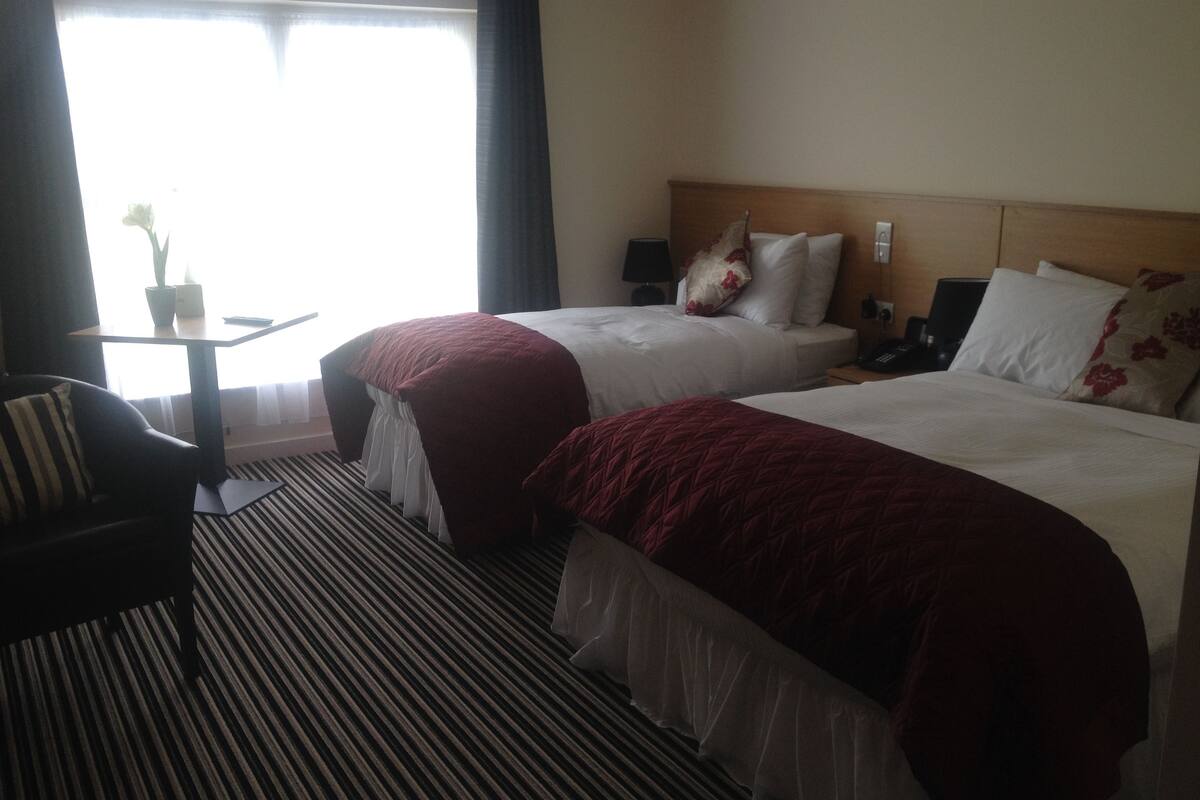 A hotel room with two beds, a bedside table, a chair, and a window with curtains.