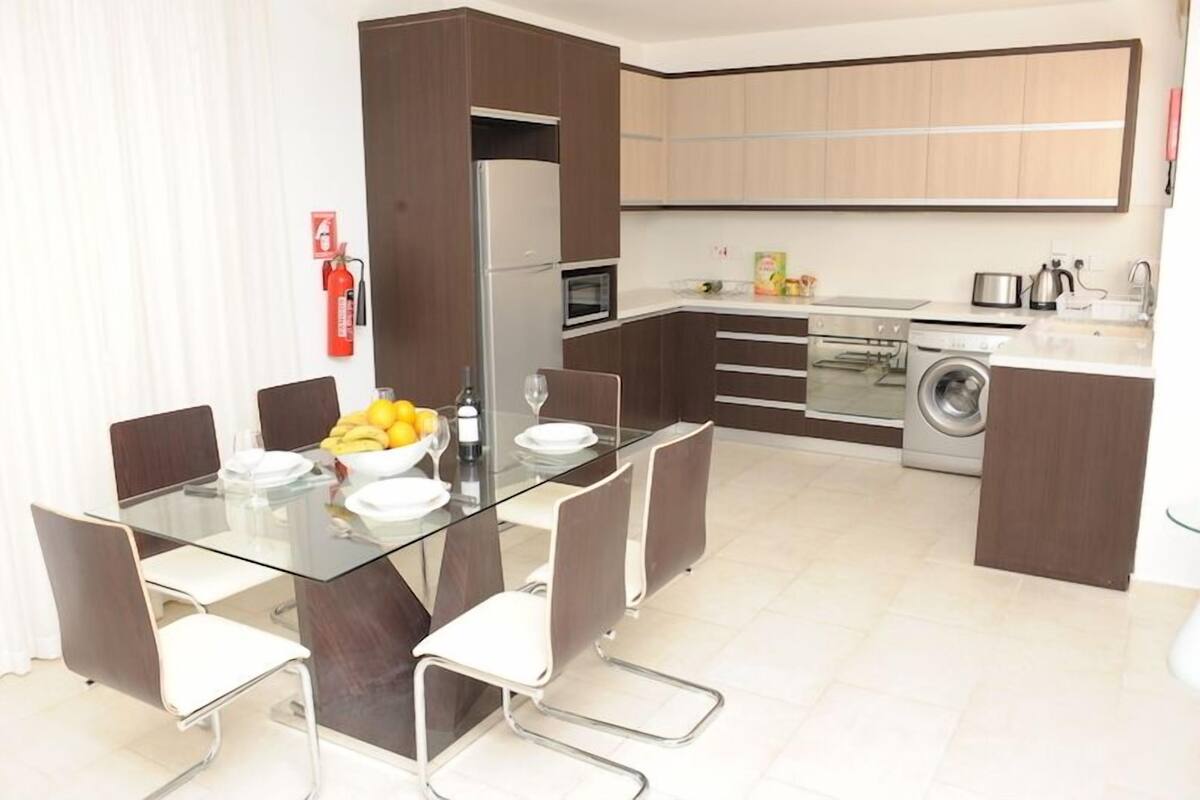 A modern kitchen with a dining area, including a glass table, chairs, a refrigerator, microwave, oven, and washing machine.