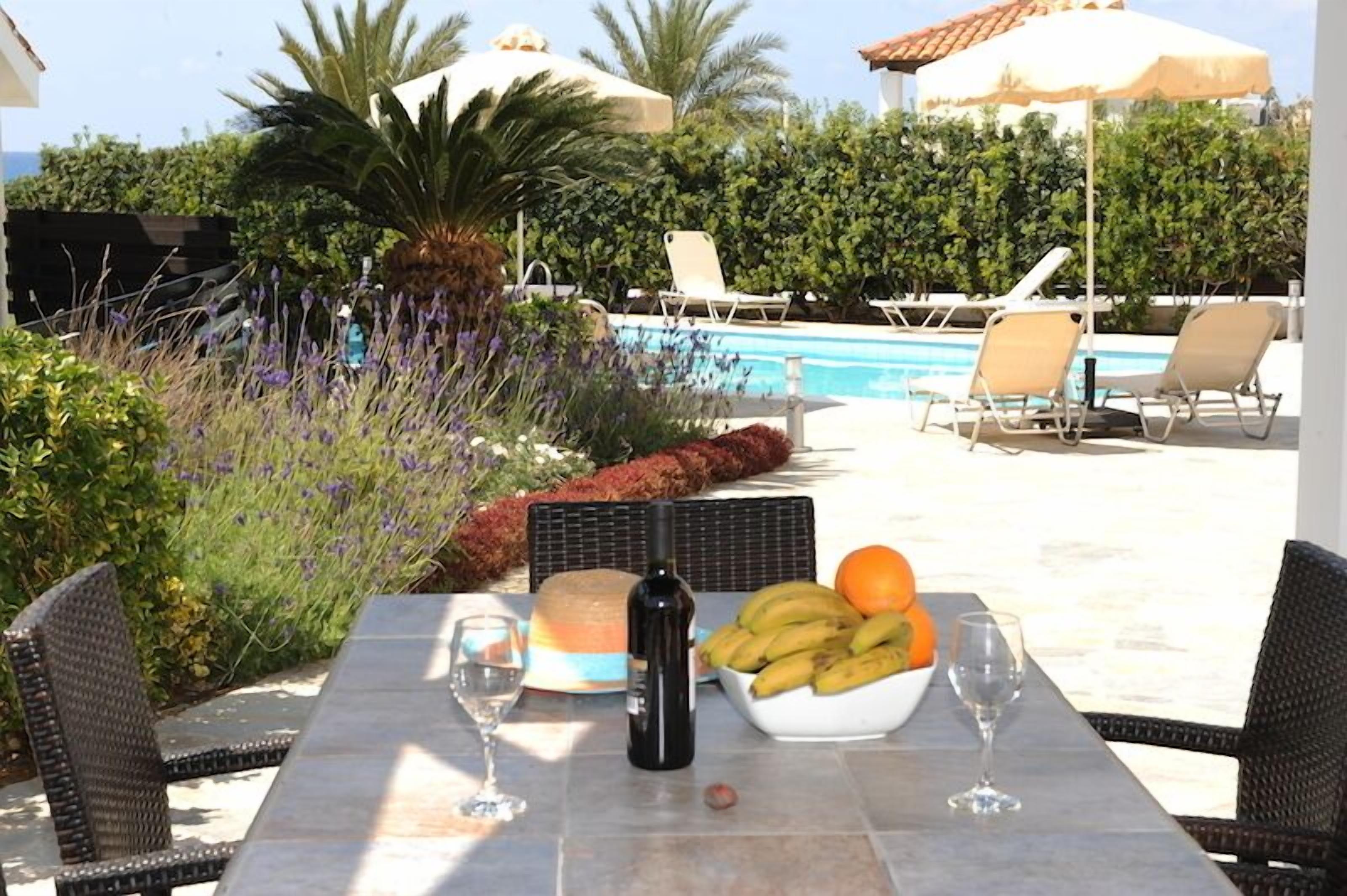Mesa à beira da piscina com uma garrafa, copos de vinho, uma cesta de frutas e uma tábua de queijos.