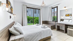 A modern hotel room with a bed, a desk, a mirror, and a view of greenery outside. - Platzia Beach Villas (Kissonerga)