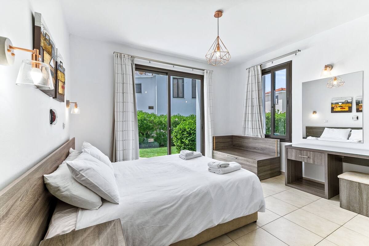 A modern hotel room with a bed, a desk, a mirror, and a view of greenery outside.