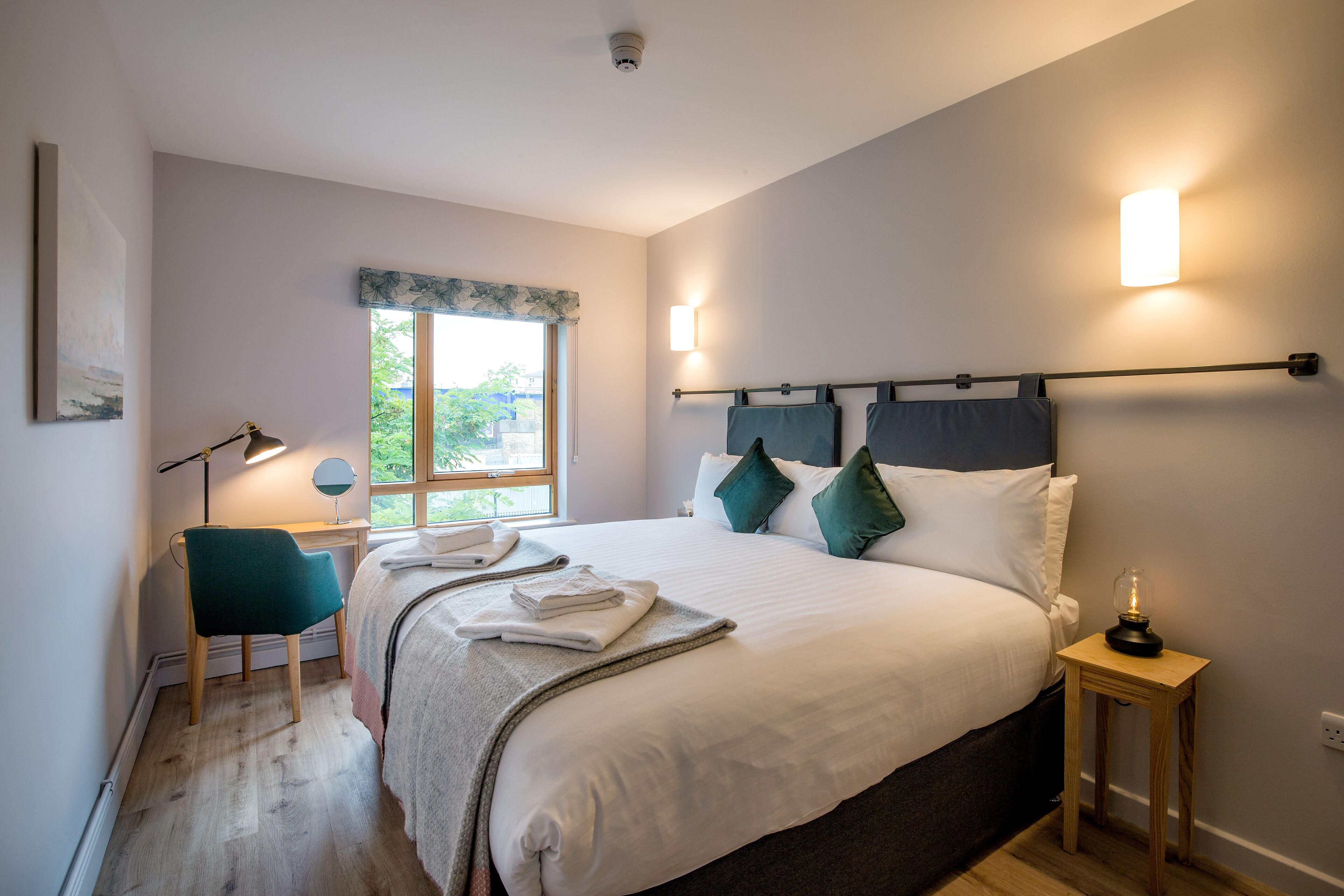 A neatly arranged bedroom with a large bed, bedside lamps, a desk, and a window with a view.