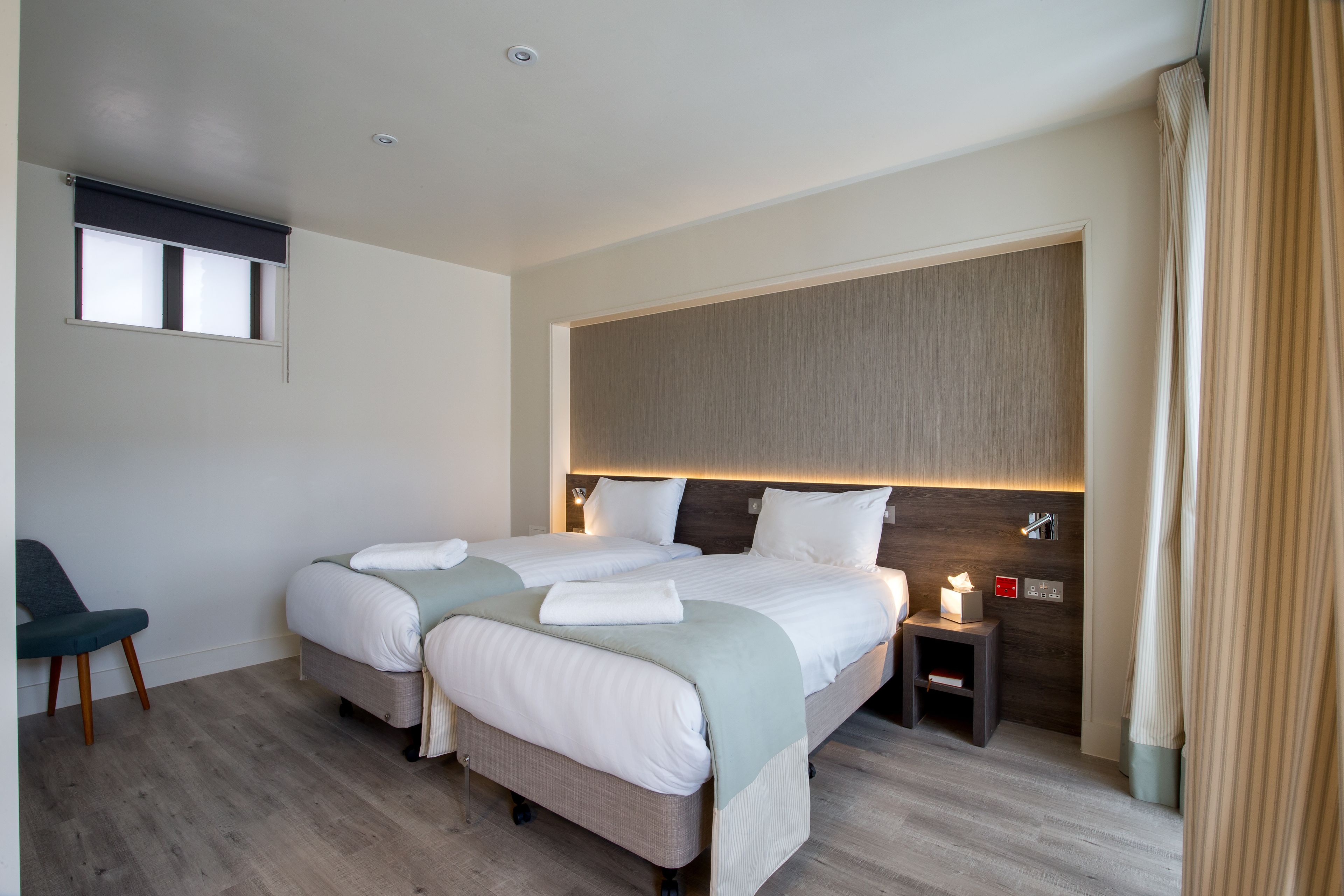 Quarto de hotel com duas camas, uma pequena mesa de cabeceira com luminária, uma cadeira e uma janela com persianas.