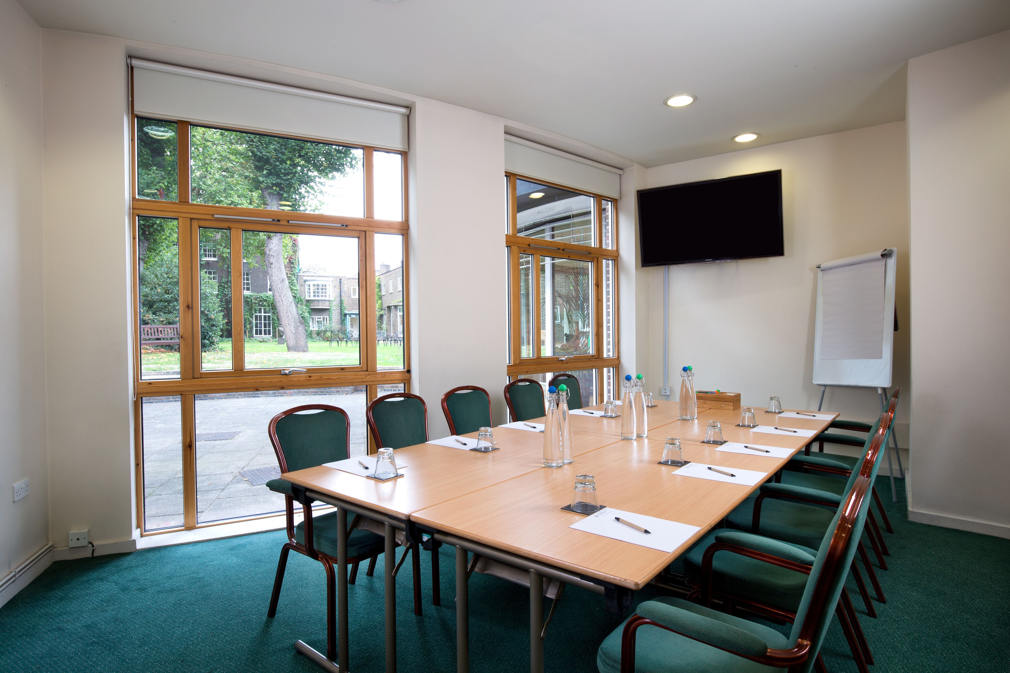 Une salle de conférence avec une longue table, des chaises vertes, un tableau blanc et un téléviseur fixé au mur.