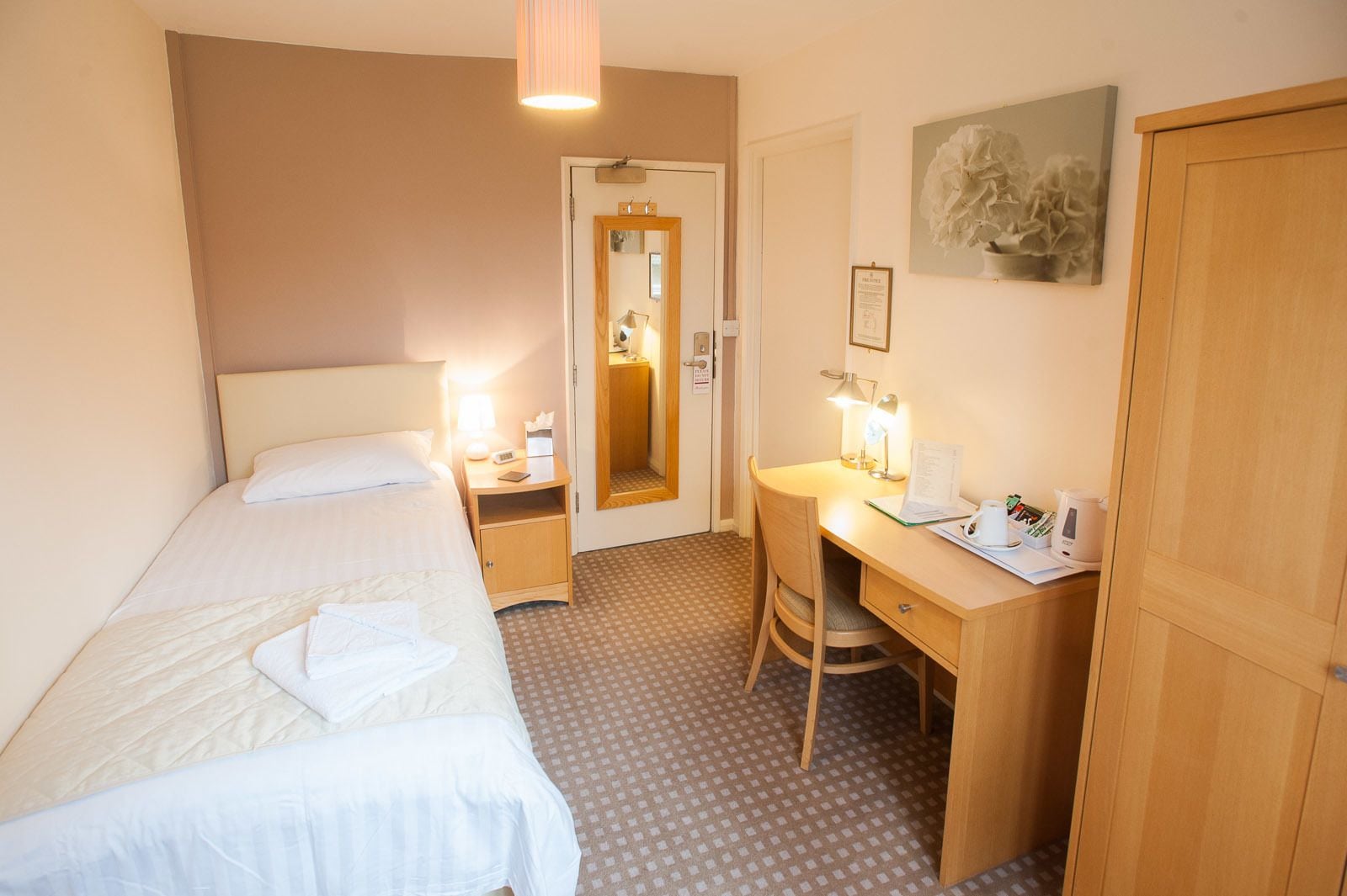 A hotel room with a bed, a desk, a chair, a wardrobe, a mirror, and a door.