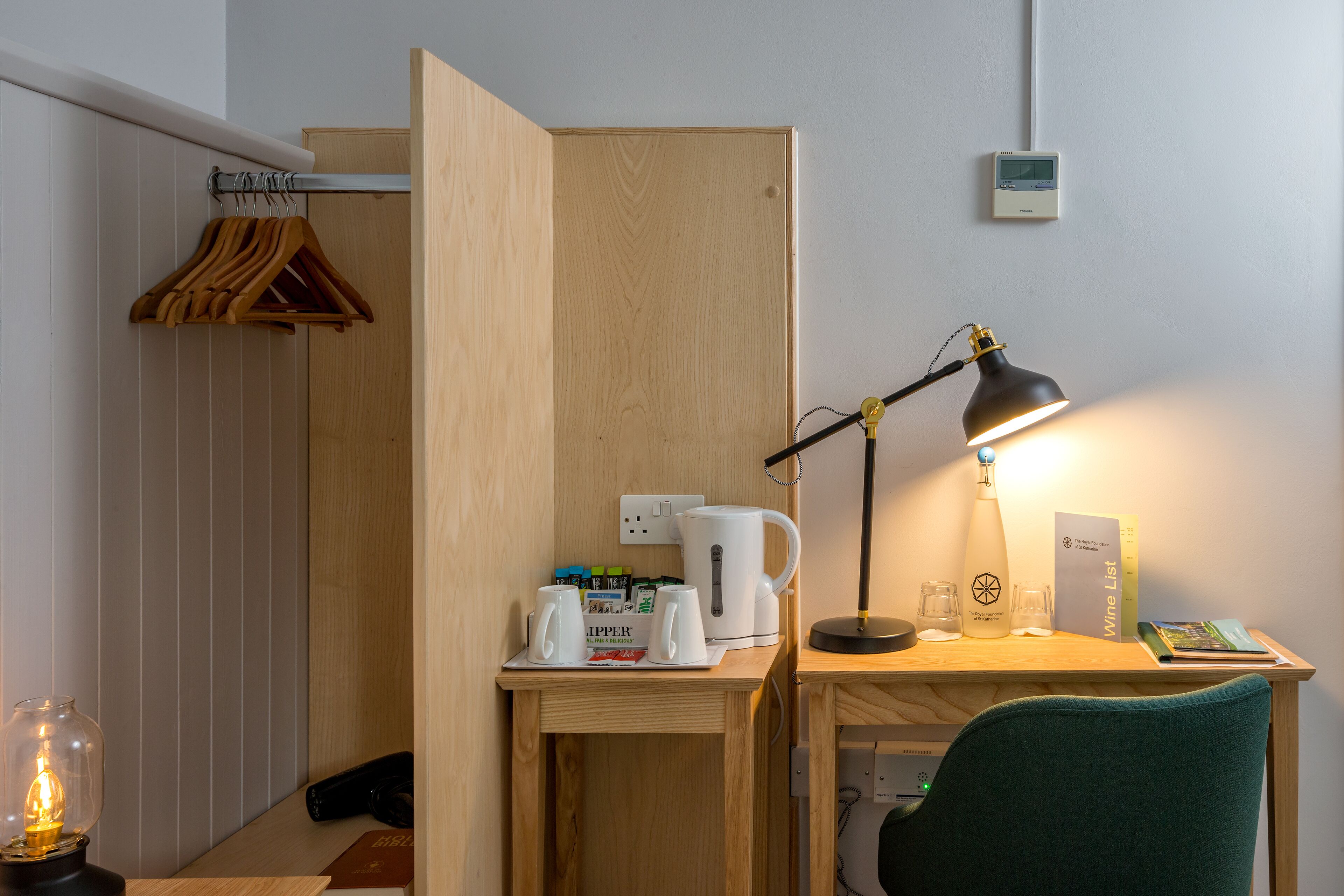 A neatly organized room with a wooden desk, a green chair, a coffee maker, a kettle, a lamp, and a thermostat on the wall.