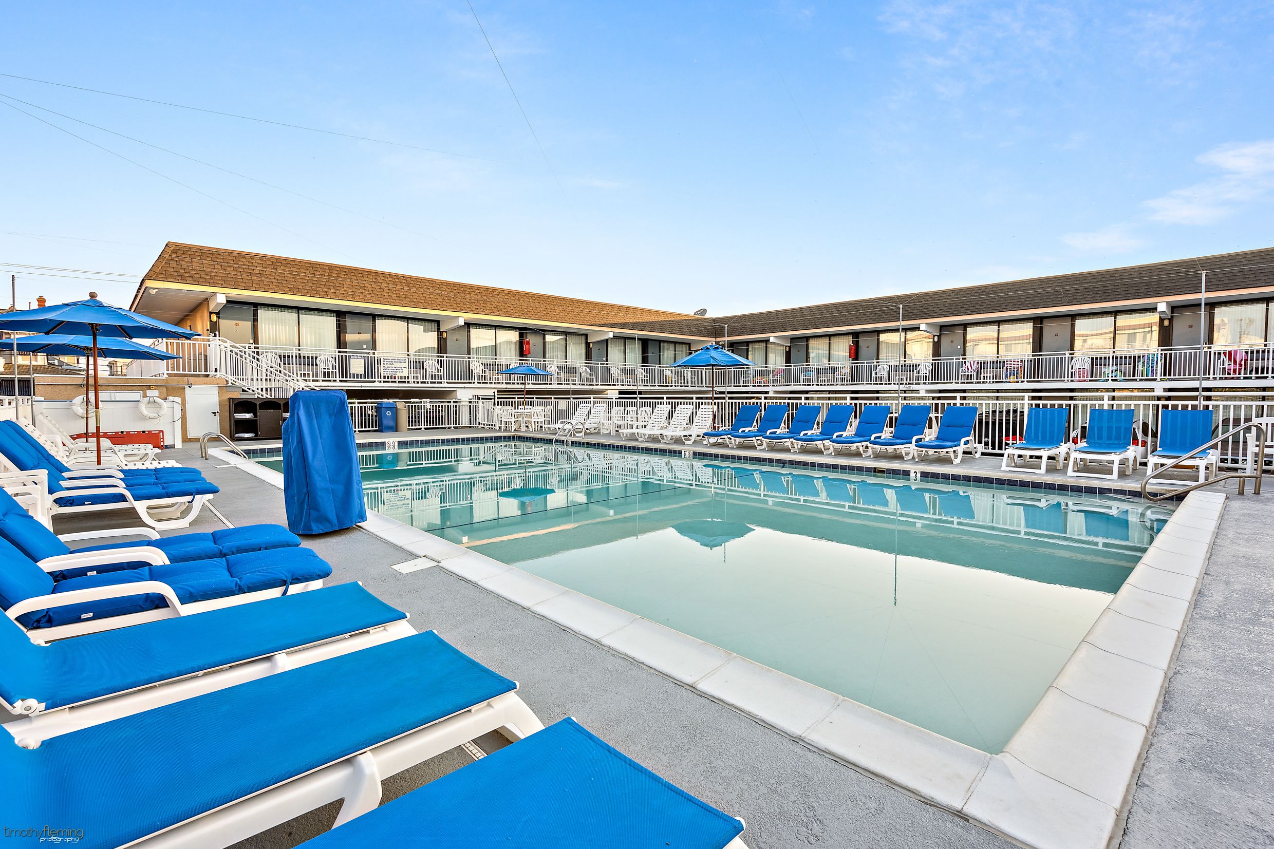A swimming pool with blue lounge chairs and umbrellas, a building with a tiled roof, and a clear sky.