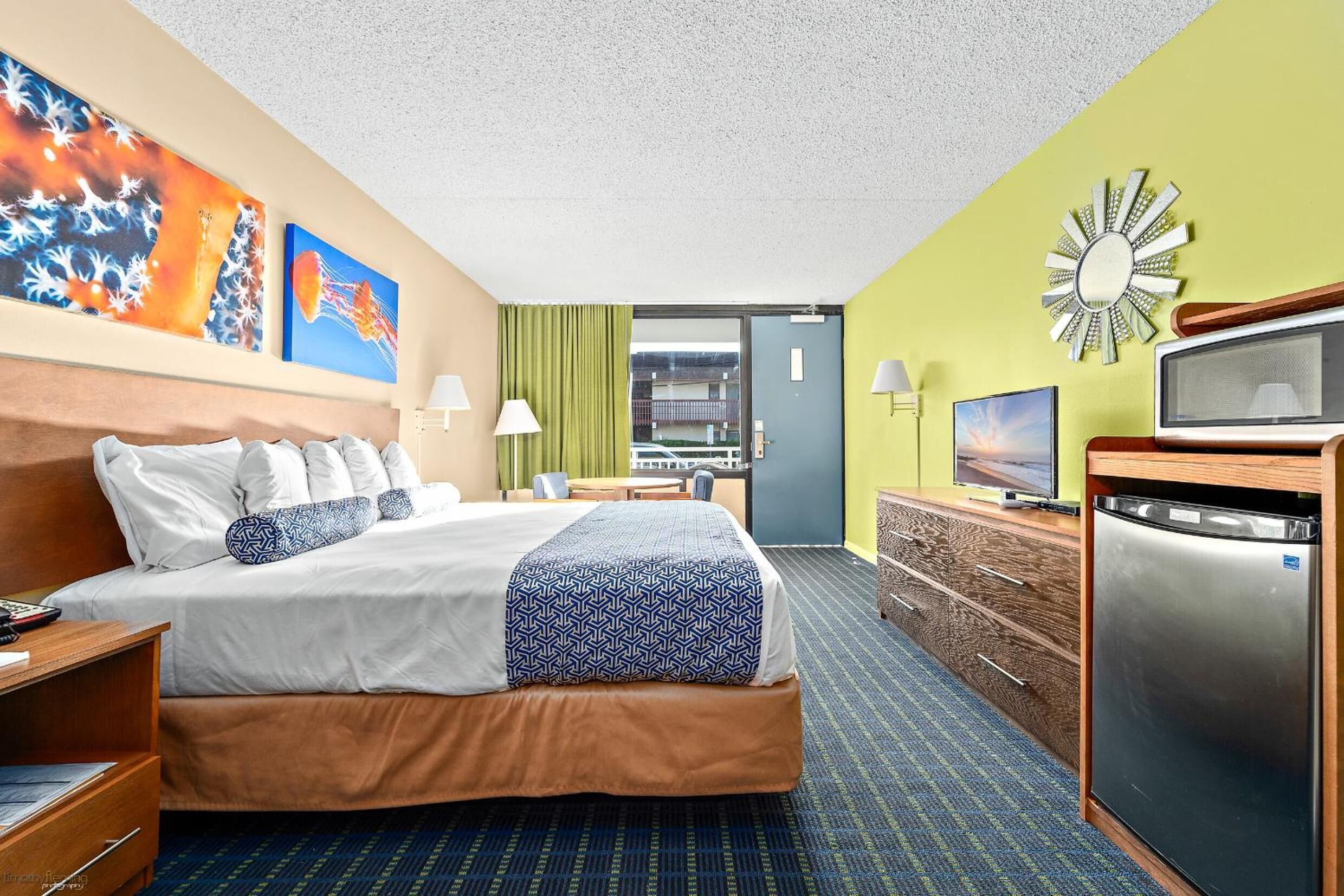 A hotel room with a bed, a television, a refrigerator, and a desk with a lamp.