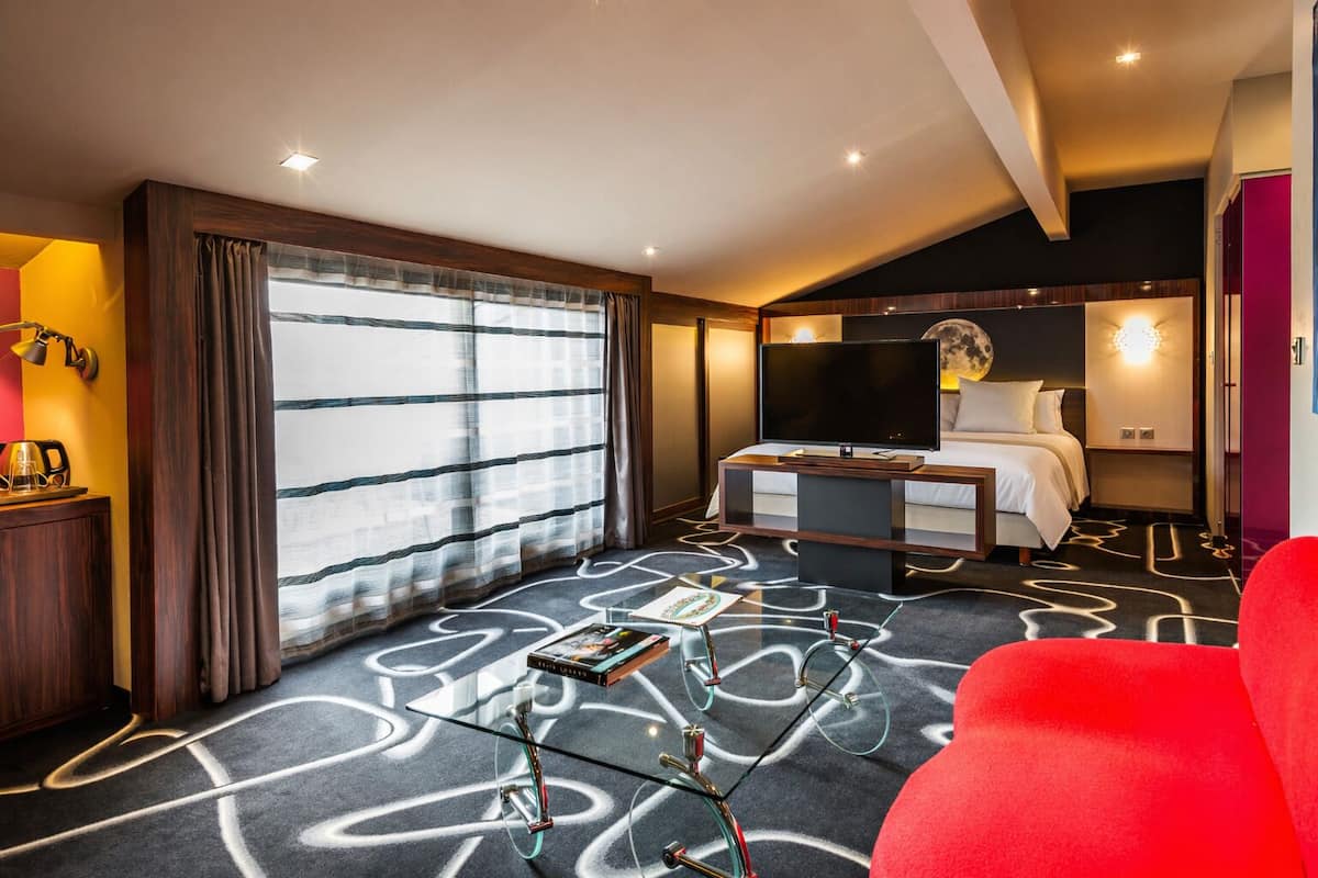 A hotel room with a bed, a desk with a TV, a red chair, and a large window with curtains.