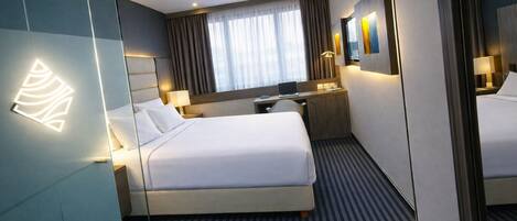 A modern hotel room with a large bed, a desk, a chair, a mirror, and a window with curtains.