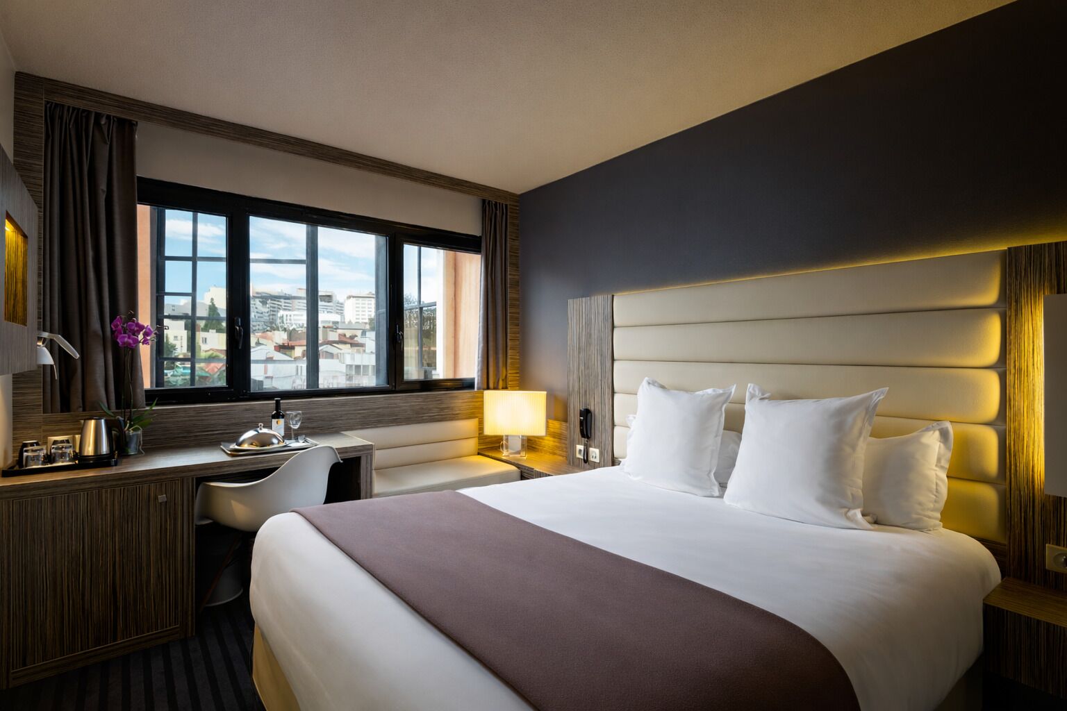 Habitación de hotel con una cama grande, un escritorio, una silla, un ventanal con vista a la ciudad y un cabecero moderno.