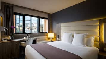 Habitación de hotel con una cama grande, un escritorio, una silla, un ventanal con vista a la ciudad y un cabecero moderno.