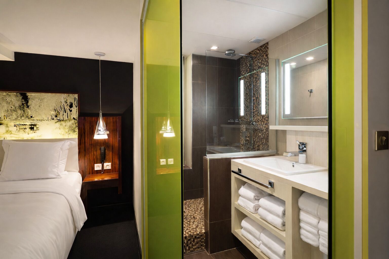 A modern hotel room with a bed, bedside lamp, and a bathroom with a sink, mirror, and towel rack.