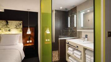A modern hotel room with a bed, bedside lamp, and a bathroom with a sink, mirror, and towel rack.