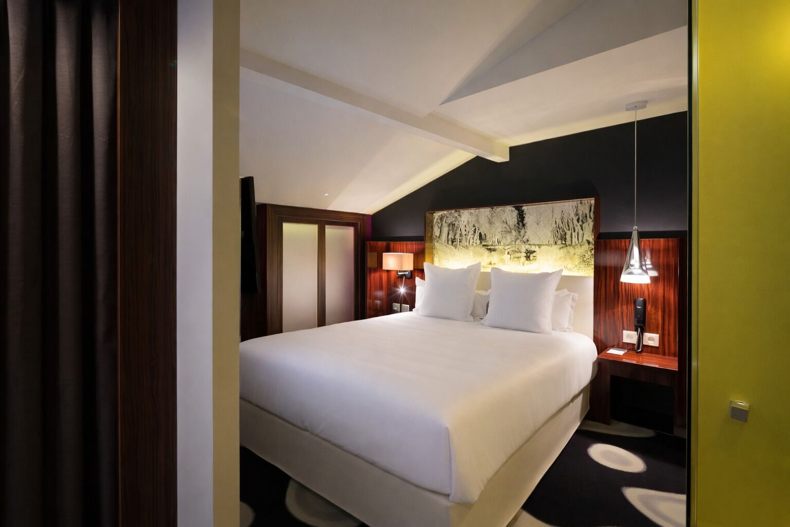 A hotel room with a large bed, a headboard with a painting, bedside lamps, and a small table with a lamp.